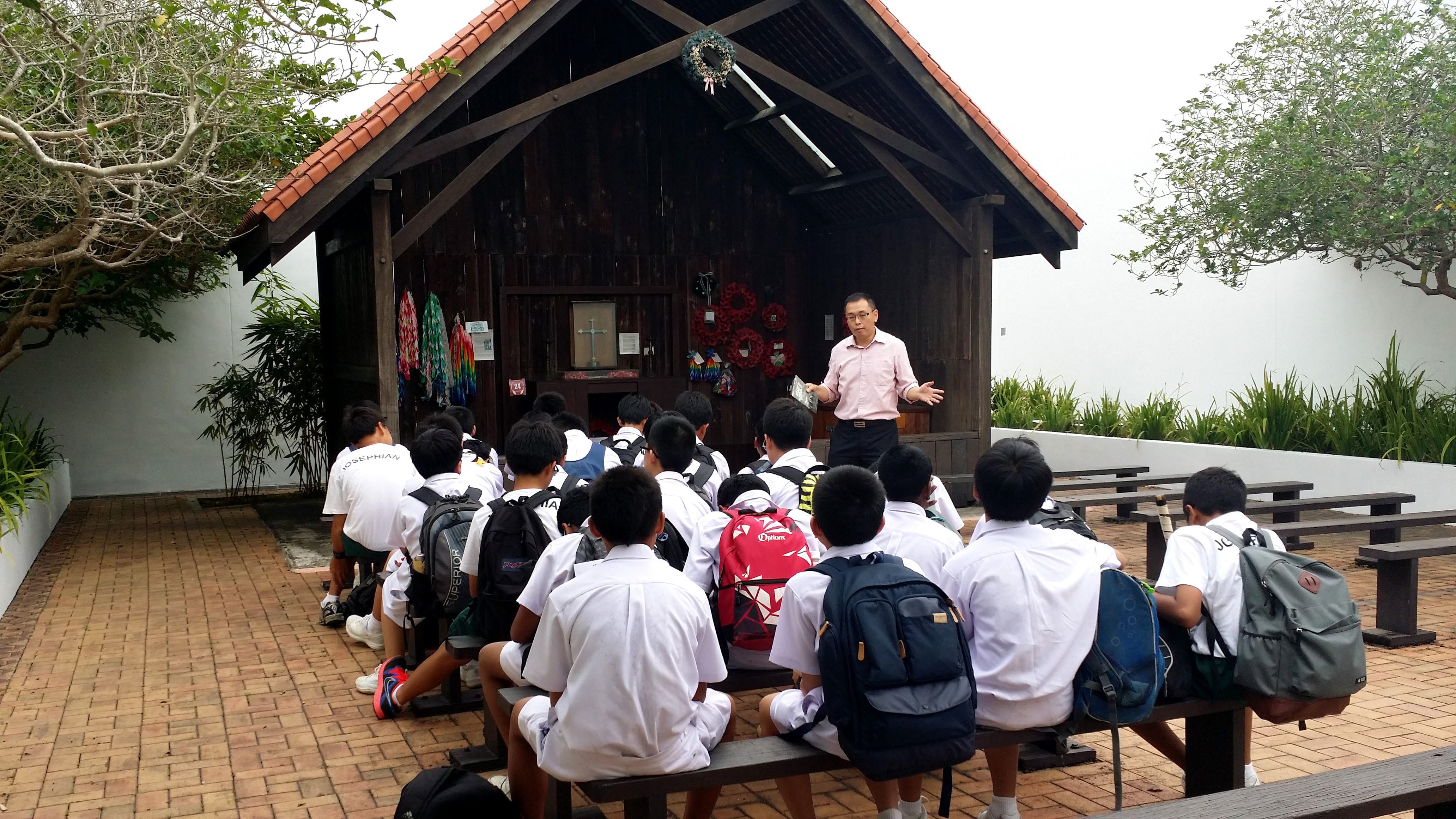 A man in a pink shirt addresses uniformed students with backpacks seated on benches outside a wooden chapel.